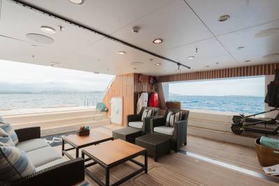 A luxurious yacht deck with comfortable seating and coffee tables in the foreground overlooks a calm blue ocean and distant islands in the background.