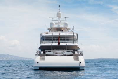A large white yacht with the name "SHAI LUXURY" on its stern is at sea with a hazy coastline in the background under a cloudy sky.