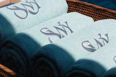 Rolled light blue towels with a dark monogram are neatly stacked in a woven basket, with dappled sunlight casting shadows across them.