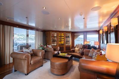 Luxurious leather seating and a large ottoman fill the foreground of a yacht's lounge, while large windows in the background offer views of the ocean and a distant coastline.