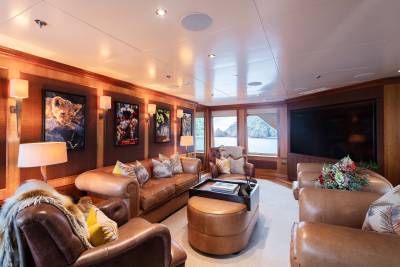 Luxurious leather seating and artwork adorn the richly paneled interior of a yacht's lounge, with a large window offering a view of a calm, rocky bay.