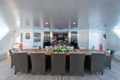 A long, polished wooden dining table set with a floral centerpiece and chairs occupies the foreground, while the background reveals an open doorway leading to a luxurious interior lounge area with a large television.