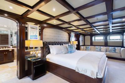 A luxurious yacht bedroom features a tufted headboard bed in the foreground and a plush seating area with windows in the background.