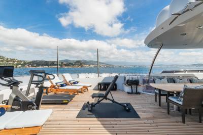 A well-equipped outdoor gym on a yacht deck faces a scenic coastline under a bright, cloudy sky.