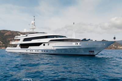 A large white yacht floats on blue water in the foreground, with a hilly, tree-covered coastline under a cloudy sky in the background.