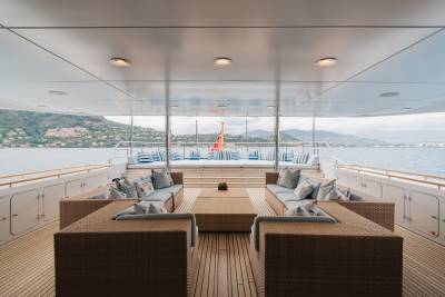 Wicker sofas and a wooden deck fill the foreground, while a calm sea and a distant, tree-covered coastline form the background.