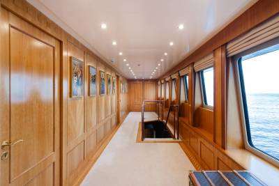 A long, wood-paneled hallway with framed pictures on the left and a large window overlooking the ocean on the right leads to a staircase going down.