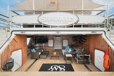 A spacious, open-air deck of a yacht, featuring comfortable seating and water sports equipment in the foreground, with the yacht's name "SHAFATY" prominently displayed on the upper deck in the background.