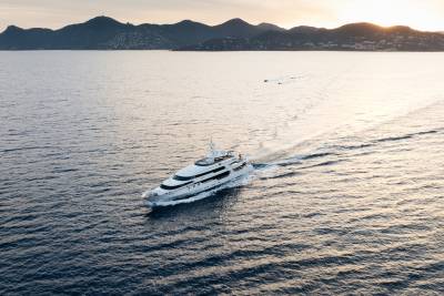 A large white yacht cruises through calm blue water, leaving a white wake, with a hazy, mountainous coastline in the background under a soft sky.