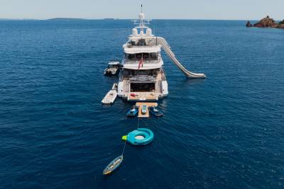 A large yacht with a waterslide and several smaller boats is anchored in the calm blue sea, with a rocky coastline visible in the distance.