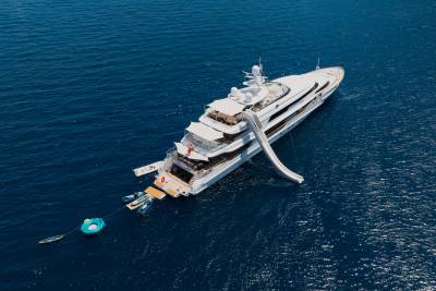 A large white yacht with a slide extending from its deck floats on the deep blue ocean, with inflatable toys tethered to its stern.