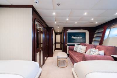 Two beds flank a plush sofa with decorative pillows in the foreground, while a hallway with wooden doors and a screen displaying a yacht at sea stretches into the background.