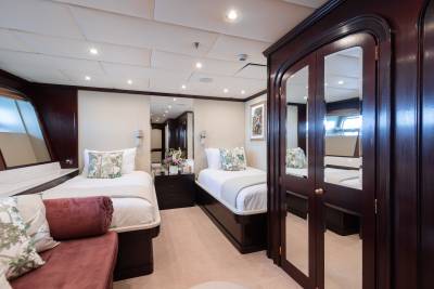 A luxurious yacht cabin features two twin beds with floral pillows in the foreground, and a large mirrored wardrobe and a window with a view of the water in the background.