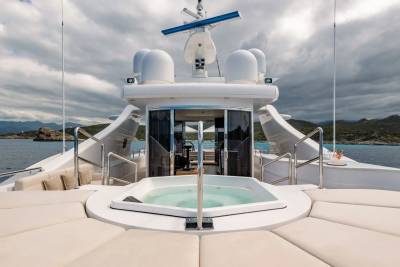 A luxurious yacht with a jacuzzi in the foreground sails on the sea with a cloudy sky and distant hills in the background.