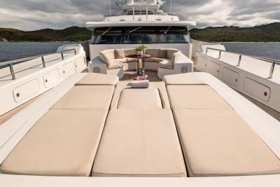 Sun loungers and a seating area on the deck of a yacht are in the foreground, with a calm sea and a tree-covered coastline in the background.