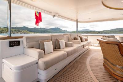 A plush, cream-colored sofa and wicker chairs furnish the deck of a yacht, with a backdrop of lush, green islands under a cloudy sky.