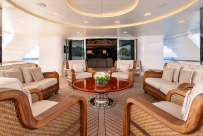 Luxurious wicker and wood seating arrangements surround a polished red table on a yacht deck, with a glimpse of a dimly lit interior and a tropical landscape visible through open glass doors in the background.