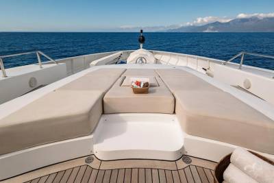 A luxurious yacht's bow with plush seating in the foreground faces a calm blue sea and distant, hazy mountains under a clear sky.