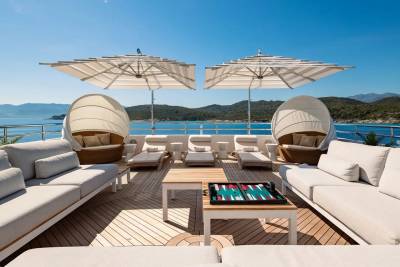 White sofas and lounge chairs with umbrellas are arranged on a wooden deck in the foreground, while a calm blue sea and green hills are visible in the background.