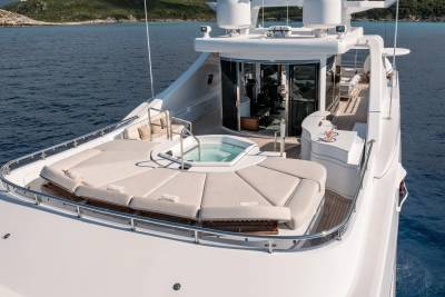 A luxurious yacht with a large sunbathing area and jacuzzi in the foreground sails on calm blue water with islands in the background.