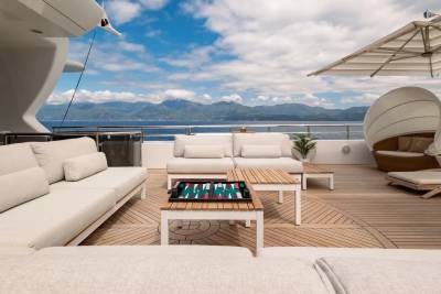 White sofas and a backgammon board are on a wooden deck in the foreground, with a serene ocean and distant mountains under a blue sky in the background.
