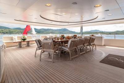 A formal dining table set for a meal on a yacht's deck is surrounded by comfortable seating, with a scenic view of islands and water in the background.