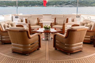 A circular arrangement of plush armchairs and a table with a floral centerpiece sits on a teak deck, with a calm sea and distant hills forming the background.