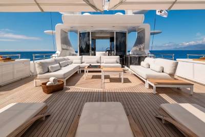 Plush white sofas and lounge chairs fill the wooden deck of a luxurious yacht, with a clear blue sky and distant, hazy mountains forming the backdrop.