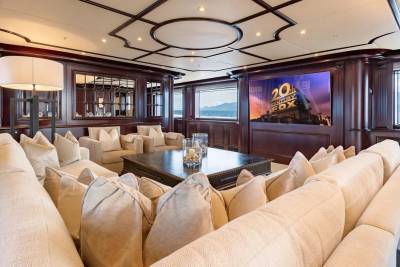 A plush, cream-colored sectional sofa fills the foreground of a luxurious yacht's entertainment room, while a large screen displaying the 20th Century Fox logo and windows with a scenic view of mountains and water form the background.