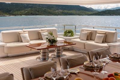 A luxurious yacht deck with comfortable seating and a dining table in the foreground is set against a backdrop of calm blue water and lush green hills.