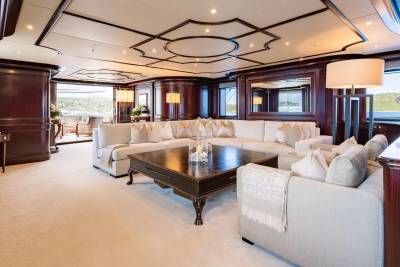 A spacious, cream-carpeted living area with a large sectional sofa and dark wood accents opens to a sunlit deck with ocean views in the background.