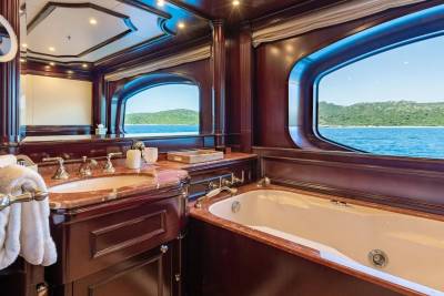 A luxurious yacht bathroom with a marble vanity and bathtub in the foreground overlooks a serene blue ocean and green hills in the background.