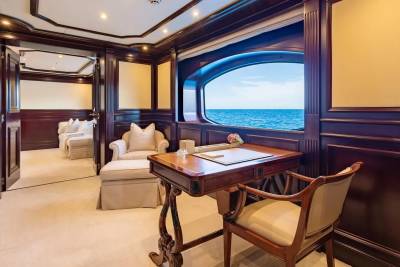 A wooden desk and chair sit in the foreground of a luxurious room with a large window overlooking the ocean, while a plush seating area is visible in the background.