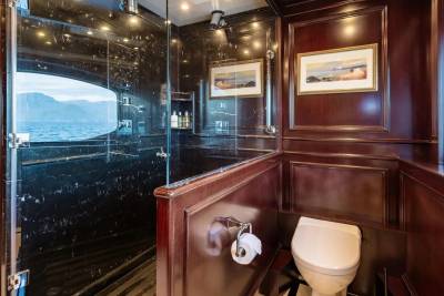 A luxurious yacht bathroom features a black marble shower with a large oval window overlooking the sea in the foreground, and a polished wood-paneled wall with framed artwork and a toilet in the background.