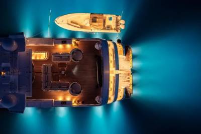 A large, illuminated yacht and a smaller tender boat float on dark blue water, with bright underwater lights creating a glowing effect.
