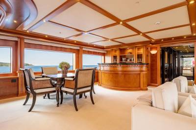A dining table with chairs sits in the foreground of a luxurious yacht's interior, with a polished wooden bar and ocean views in the background.