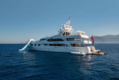 A large white yacht with a slide is anchored in calm blue water, with hazy mountains in the background under a clear sky.