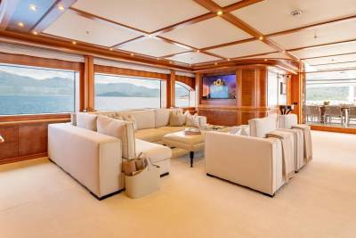 A spacious, cream-colored sectional sofa and armchairs fill the foreground of a luxurious yacht's lounge, while large windows in the background reveal a serene seascape with distant mountains and an outdoor dining area.