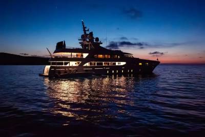 A luxurious yacht with illuminated windows floats on calm water in the foreground, while a dark, hilly coastline is visible against a twilight sky in the background.