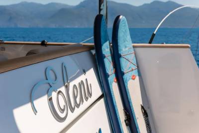 Two blue paddleboards lean against the side of a white yacht named "Eleni" with a calm blue ocean and distant mountains in the background.