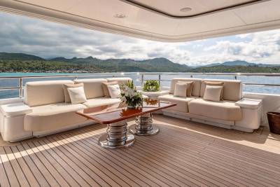 Plush sofas and a coffee table sit on a wooden deck overlooking a serene bay with tree-covered hills under a cloudy sky.