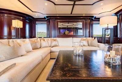 A plush sectional sofa and decorative glass vases sit on a dark coffee table in the foreground, while a richly paneled room with large windows revealing a scenic landscape forms the background.