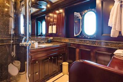 A luxurious yacht bathroom features a marble shower stall in the foreground and a dark wood vanity with oval windows in the background.