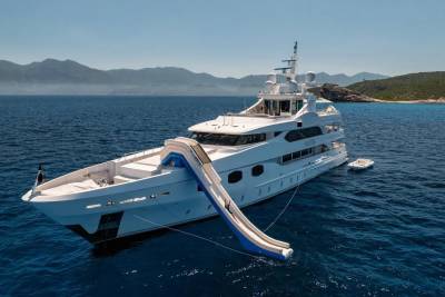 A large white yacht with a blue slide extending from its deck floats on calm blue water, with a mountainous coastline visible in the distance under a clear sky.