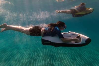 A woman glides through clear blue water on a sea scooter in the foreground, while another person on a similar device swims in the sandy background.