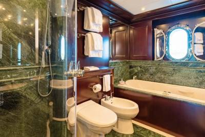 A luxurious bathroom with a glass-enclosed shower and bidet in the foreground, and a bathtub and ornate oval window in the background.