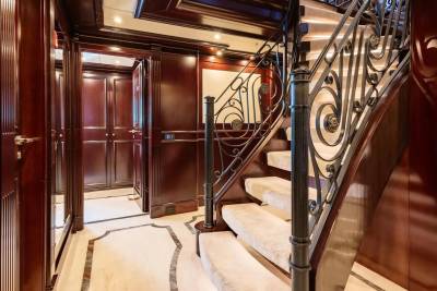 A luxurious, ornate staircase with plush carpeted steps and intricate metal railings dominates the foreground, while the background reveals a polished wood hallway with mirrored doors and elegant cabinetry.