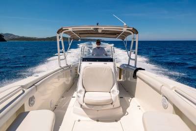 A person drives a white boat across a bright blue ocean, leaving a white wake, with a rocky island visible in the distance under a clear sky.