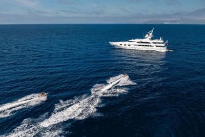 Two jet skis race across the deep blue ocean in the foreground, leaving white wakes, while a large white yacht sails in the background under a clear sky.