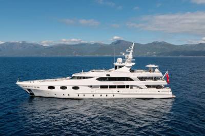 A large white yacht floats on calm blue water in the foreground, with a backdrop of distant, hazy mountains under a clear blue sky.